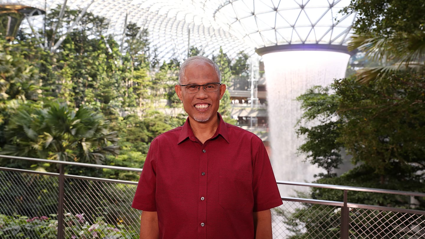 Person wearing glasses smiles in front of a large indoor waterfall and trees.